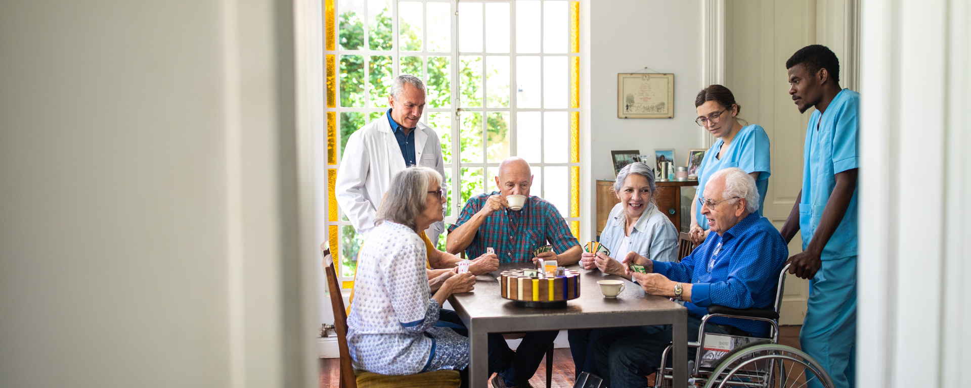 Several elderly people are sat at a dining table drinking coffee and playing cards. Three medical staff are stood around the table, engaged in conversation with those sat down.