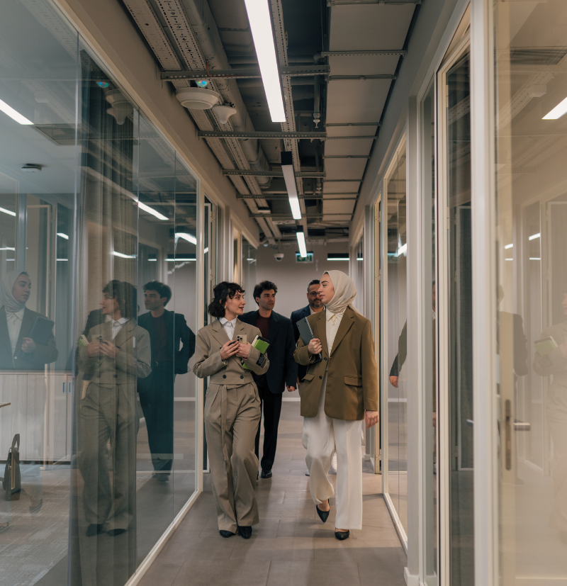 Two coworkers are walking down a hallway, surrounded by glass-fronted meeting rooms.