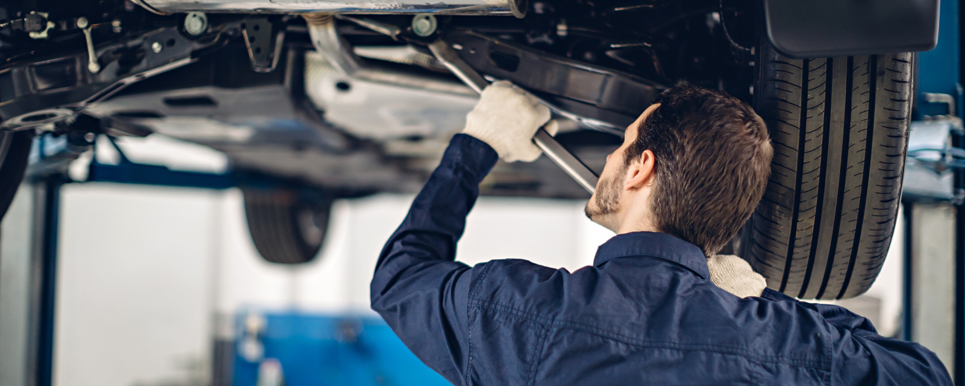 A car is elevated in a workshop and a mechanic is working on the underside.